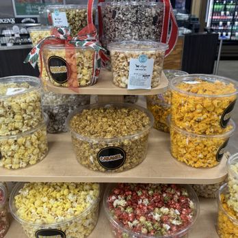 Kroger - Popcorn - Multiple containers of flavored popcorn are arranged on a tiered wooden display inside a grocery store. The popcorn flavors include caramel, cheese, and other varieties, with some containers stacked. The background shows store shelves and refrigerated sections, indicating an indoor retail environment. - Grocery Near Me - Fort Wayne, Indiana