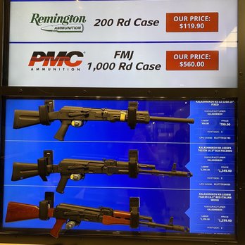 The Range at Austin - The image shows three rifles mounted on a blue background, each with a different design and stock color, along with detailed specifications and prices. The top rifle has a black stock, the middle has a black stock, and the bottom has a brown stock. Above the rifles, a digital display advertises ammunition prices for a shooting range or firearm store, including Remington and PMC brands, with prices for different case sizes. The setting appears to be an indoor retail or shooting range environment. - Guns & Ammo Near Me - Austin, Texas