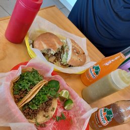 Tacos & Torta - A meal with tacos and a sandwich is served on a wooden table, accompanied by condiments including hot sauce, mayonnaise, and a drink.