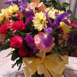 A vibrant flower arrangement featuring irises, daisies, carnations, and other flowers, decorated with a gold ribbon, placed in a vase on a table.