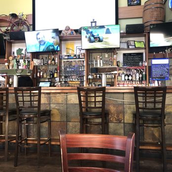 Firehouse Pizza & Pub - Inside/bar area - The photo shows the interior of Firehouse Pizza & Pub, with a bar counter lined with wooden high chairs. Behind the bar, there are several TV screens displaying various programs, including a news broadcast, a racing game, and a person in a green jacket. The bar area has bottles of alcohol, glassware, and a chalkboard menu. Decorative elements include a mounted bicycle on the wall and various small items on shelves. The setting appears casual and suitable for socializing and watching sports. - Pizza Near Me - Normal, Illinois