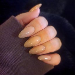A hand with manicured nails painted in a nude shade with gold glitter accents along the edges, resting on a dark fabric background.