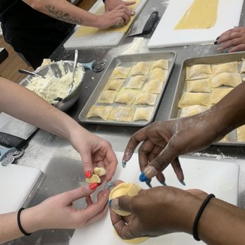 Selfup - Hand folded home made from scratch - Multiple individuals are engaged in a cooking activity, preparing homemade pasta or ravioli. One person is filling and sealing dough, while others are rolling out dough and assembling pasta on trays. The workspace includes cutting boards, trays with pasta, and a bowl of filling, indicating a hands-on culinary class focused on pasta making. - Cooking Classes Near Me - Boston, Massachusetts