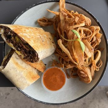 Buen Provecho - Brunch burrito at Buen Provecho. - A plate featuring a sliced burrito with beef filling, fried plantains, and a small cup of red sauce, served on a black and white dish. - Latin American Near Me - Albuquerque, New Mexico