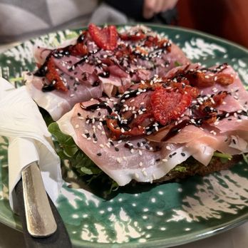 Criterion coffee - Prosciutto Toast - A close-up of a prosciutto toast featuring thin slices of prosciutto, fresh strawberries, sesame seeds, and a drizzle of balsamic glaze, served on a green patterned plate. The dish is garnished with leafy greens underneath, and a knife wrapped in a napkin rests on the side of the plate. The background shows part of a person's arm and a brown chair. - Coffee & Tea Near Me - Pasadena, California