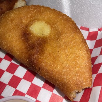 Sabe a zulia - Shredded beef empanada - A large, fried shredded beef empanada with a crispy, golden-brown crust rests on red and white checkered paper. The empanada has a rounded, semi-circular shape with a slightly uneven surface, indicating a flaky, fried dough exterior. Part of another food item and a small container of dipping sauce are visible nearby, suggesting a casual dining setting. The background includes a piece of foam or packaging material. - Venezuelan Near Me - Chicago, Illinois