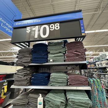 Walmart Supercenter - Multiple shelves hold neatly folded towels and blankets in various colors, including gray, blue, green, and maroon. A large digital price sign above shows the price as $10.98. The store has a high ceiling with visible metal beams, and other shelves with additional products are visible in the background. A shopping cart is partially visible on the right side. - Grocery Near Me - Dixon, Illinois
