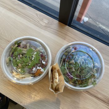 CAVA - to-go bowls with complimentary pita - Two plastic to-go salad bowls with clear lids and a piece of flatbread are placed on a wooden table near a window, with a sidewalk visible outside. - Mediterranean Near Me - Arvada, Colorado