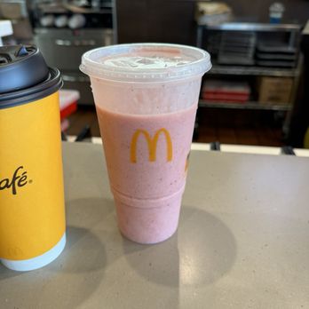 McDonald's - A pink McDonald's smoothie in a clear plastic cup with a lid is positioned on a beige counter. The cup features the yellow McDonald's logo. To the left, there is a yellow McDonald's coffee cup with a black lid. The background shows a busy restaurant interior with staff and kitchen equipment, slightly out of focus. - Fast Food Near Me - Raton, New Mexico