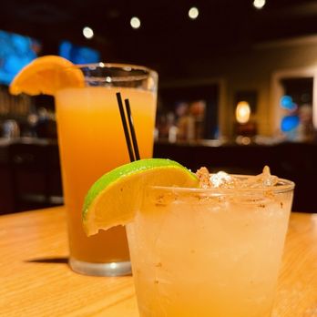 Chili's - Two drinks with lime and orange garnishes are placed on a wooden table inside a dimly lit bar or restaurant. - American Near Me - Temple, Texas
