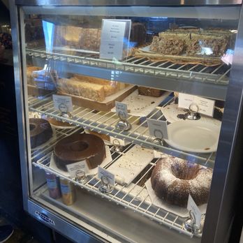 Raging Sage - The image shows a glass display case filled with baked goods such as cakes, slices, and a bundt cake, arranged on multiple shelves. Price tags are visible for some items, indicating prices like $2.50, $4.00, and $7.25. The case is situated on a wooden floor, and the background suggests an indoor bakery or cafe setting. - Coffee & Tea Near Me - Tucson, Arizona