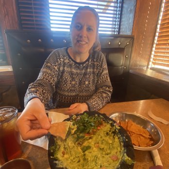 Plaza Azteca - The guacamole bowl. A must have! They make the whole thing in front of you! - A woman seated at a restaurant table reaches for a bowl of guacamole, with chips and drinks visible on the table, and wooden blinds in the background. - Mexican Near Me - Reading, Pennsylvania