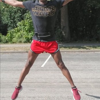 Linne Woods - A person is captured mid-air with arms and legs extended outward, outdoors on a paved surface. They are wearing a black Star Wars t-shirt, red athletic shorts, pink sneakers, sunglasses, and a cap. The background features green trees and a sidewalk, indicating a park or outdoor recreational area. - Hiking Near Me - Morton Grove, Illinois