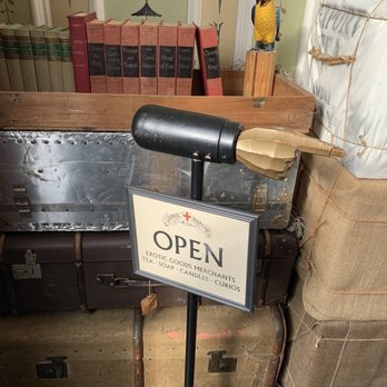 Diehl Marcus & Company - A sign reading 'OPEN' for exotic goods merchants, tea, soap, candles, and curios, is displayed on a vintage black lamp stand with a damaged lampshade, in front of antique trunks and a bookshelf. - Home Decor Near Me - Salem, Massachusetts