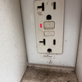 Pristine Inspections & Testing - A standard electrical outlet with two power sockets, positioned on a wall close to the floor. The outlet includes reset and test buttons, with a small red indicator light. The surrounding area shows dirt and dust, and the outlet is mounted on a white wall with some marks and grime. A label below the outlet indicates it is a protected outlet. - Home Inspectors Near Me - Champaign, Illinois