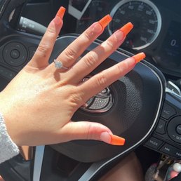Nails - A hand with long, orange acrylic nails and a diamond ring is placed on a car's steering wheel, with a photo of a person visible on the dashboard.