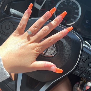 Nail Model - Nails - A hand with long, orange acrylic nails and a diamond ring is placed on a car's steering wheel, with a photo of a person visible on the dashboard. - Nail Salons Near Me - Gardendale, Alabama