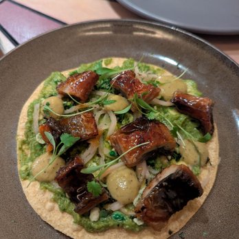 Mariscos San Pedro - Bbq eel tostada. Party in your mouth.. so so good - A seafood tostada features grilled eel pieces, garnished with microgreens, herbs, and sauce, served on a crispy tortilla. The dish is presented on a dark ceramic plate, with a colorful tiled surface and a gray plate visible in the background, indicating a restaurant setting. - Mexican Near Me - Chicago, Illinois