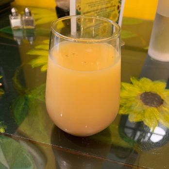 Sunflower Cafe - Mimosa! - A clear glass filled with orange juice is positioned on a table decorated with sunflower-themed artwork. The table has a dark surface with sunflower images, and the background includes a yellow wall, a menu, and a water glass. The setting appears to be a cafe or restaurant environment. - Cafes Near Me - Cooperstown, New York