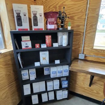 Addison Coffee Roasters - Display inside store - A black shelf holds coffee brewing devices, coffee bags, and informational cards, situated in a store with wooden walls and a window. - Coffee & Tea Near Me - Forney, Texas