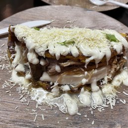 Chu... - A plate presenting a traditional Venezuelan dish with shredded chicken, melted cheese, and a creamy white sauce, garnished with small herbs. The dish is served on a round wooden plate, with some cheese sprinkled around the edges. The presentation emphasizes the layered ingredients and the rustic serving style.