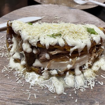 Manny's Cabin - Chu... - A plate presenting a traditional Venezuelan dish with shredded chicken, melted cheese, and a creamy white sauce, garnished with small herbs. The dish is served on a round wooden plate, with some cheese sprinkled around the edges. The presentation emphasizes the layered ingredients and the rustic serving style. - Venezuelan Near Me - Florence, Kentucky