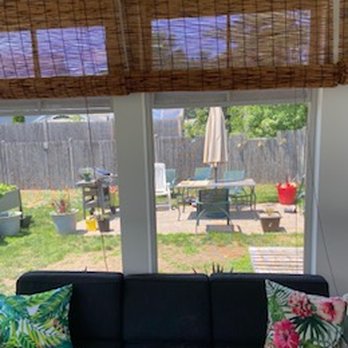 Drohan Development - The image shows a view through glass sliding doors into a backyard. Outside, there is patio furniture including a table with an umbrella, chairs, and various potted plants. A wooden fence encloses the yard, and there are trees and greenery in the background. Inside, a black sofa with colorful floral pillows is partially visible in the foreground. The scene suggests a residential outdoor space with a seating area. - General Contractors Near Me