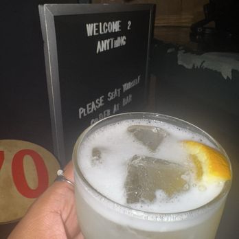 Anything - A hand holds a glass of a cold drink with ice cubes and a lemon slice, in front of a bar sign that says 'Welcome 2 Anything.' - Pizza Near Me - Brooklyn, New York