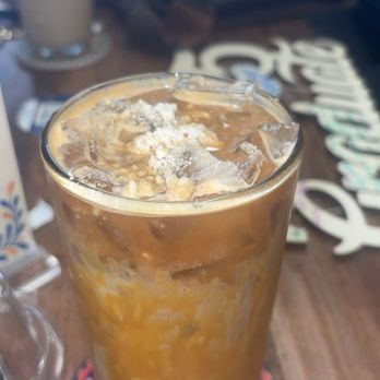 Cafe es Amor - this is the marzipan latte (i spelt it wrong in the photo ) - A tall glass of iced coffee with cream, ice cubes, and a sprinkle of powdered topping, placed on a colorful coaster on a wooden table. - Coffee & Tea Near Me - Albuquerque, New Mexico