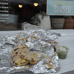 A partially eaten burrito wrapped in aluminum foil with side containers of green and red sauces on a white outdoor surface.