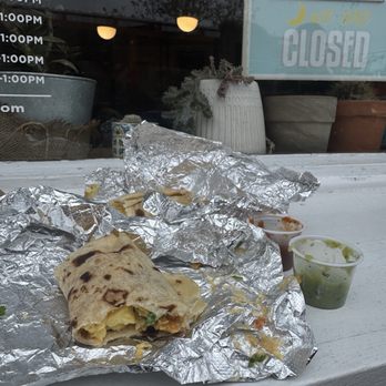 La Tejana - A partially eaten burrito wrapped in aluminum foil with side containers of green and red sauces on a white outdoor surface. - Tex-Mex Near Me - Washington, DC, District of Columbia