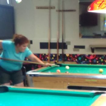 Silva Lanes-Eucan Bowl - My daughter taking her shot.  It was her first time playing pool. - A woman with brown hair tied back, wearing a turquoise shirt, is leaning over a pool table, aiming her cue stick at the balls. The setting is an indoor recreational space with multiple pool tables, cue racks, and a television mounted on the wall. The background includes a colorful patterned wall and a neon sign, indicating a casual entertainment venue. - Bowling Near Me - Albuquerque, New Mexico