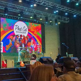 Credence Clearwater Revisited tonight - A stage features musical instruments such as guitars, a drum set, and microphones, with a large digital screen displaying the text 'Phat Cat Swinger' and colorful graphics. Audience members are seated in front of the stage, and stage lighting illuminates the scene. The venue appears to be an indoor performance space with a professional setup.