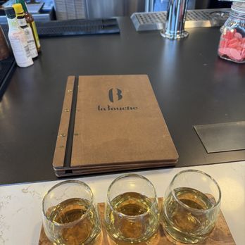 La Bouche - French white flight - Three glasses of white wine are placed on a wooden tray at a bar counter in a restaurant or wine bar setting. - Wine Bars Near Me - Denver, Colorado