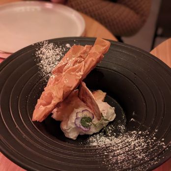 Prime Bar - Chicago - Lemon Tart - A plated dessert featuring a layered pastry, whipped cream, and a decorative edible flower, presented on a black plate with powdered sugar. - Cocktail Bars Near Me - Chicago, Illinois
