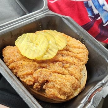Wingstop - Hot Honey Rub chicken sammich - A fried chicken sandwich topped with pickles is served in a black foam container, held by a person wearing a red shirt. - Chicken Wings Near Me - Fairfax, Virginia