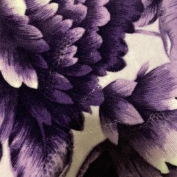The image shows a close-up of fabric with a floral pattern in shades of purple and black. The design includes detailed petals and leaves with textured, layered appearances, highlighting the fabric's intricate print and rich color contrast.