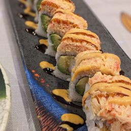 Yummy Roll - Sushi rolls with avocado and seafood filling are arranged in a line on a black rectangular plate. The rolls are topped with drizzles of yellow and dark sauces, likely mayonnaise and eel sauce. The plate is placed on a light-colored tablecloth, and the sushi presentation emphasizes colorful ingredients and garnishes.