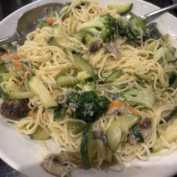 Clara's - Angel hair pasta with vegetable primavera sauce - A plate of vegetable primavera pasta includes cooked noodles mixed with broccoli, zucchini, and mushrooms. The dish appears to be served hot, with visible steam and a light sauce coating the vegetables and noodles. The presentation is simple, on a white round plate, with a metal fork resting on the edge. The background suggests a restaurant setting. - Italian Near Me - Woodridge, Illinois