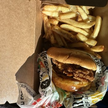 Peri Peri Original - Chicken Sandwich & Fries - A fried chicken sandwich with a bun, fried chicken fillet, and sauce, served alongside a portion of French fries in a cardboard takeout box. The sandwich is partially wrapped in branded paper, and the fries are golden and crispy. The container is open, revealing the food inside, with a green fabric visible on the right side of the image. - Chicken Shop Near Me - Laurel, Maryland