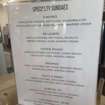 Half Baked Cafe - Sundaes too! - A clear plastic menu board features a list of specialty sundaes offered at a dessert shop. Each sundae includes specific ingredients such as custard, fudge, marshmallow, chocolate chips, whipped cream, and other toppings. The menu provides detailed descriptions for each flavor, including S'mores, PB Lovers, Cookie Dough, Brownie, Salty & Sweet, Almond Joy, and Kids Kreation, with prices listed for the Kids Kreation. The background shows part of the shop interior with some lighting and shelving. - Ice Cream & Frozen Yogurt Near Me - Lockport, New York