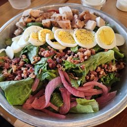 A large, mixed salad in a metal bowl includes sliced boiled eggs topped with black pepper, chopped bacon, grilled chicken pieces, and pickled red onions. The salad is placed on a wooden table with small containers of dressing or sauces visible in the background. The ingredients are arranged in sections, showcasing a variety of textures and colors.