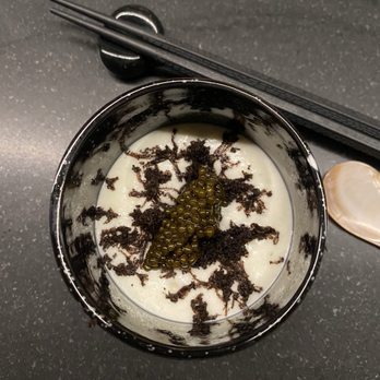 Sho - Chawanmushu- truffled dashi, cauliflower espuma, osetra caviar aka umami bomb - A bowl of chawanmushi, a traditional Japanese savory egg custard dish, is shown on a dark, textured table. The dish is garnished with black truffle shavings and a small cluster of osetra caviar in the center. Chopsticks rest on a holder above the bowl, and a spoon is placed to the side. The presentation emphasizes the luxurious ingredients used in the dish. - Japanese Near Me - Chicago, Illinois