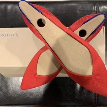 Rothy's - Red pointed shoes with beige soles and blue trim are positioned on top of a Rothy's box. The shoes are made of a fabric material, with a sleek, pointed toe design. The soles are smooth and beige, with two circular holes near the heel area. The shoes are placed on a black surface, likely a table or bench, with the box serving as a base. The box has the brand name 'ROTHY'S' printed on it. - Shoe Stores Near Me - Washington, DC, District of Columbia
