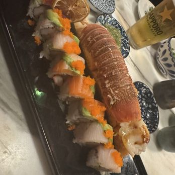 The 698 - Grubbings - The image shows a black rectangular platter with assorted sushi rolls, including fish, avocado, and orange fish roe, arranged in a line. Next to the sushi, there is a whole cooked fish garnished with lemon slices and wasabi. The table has various small plates, bowls, and a glass of beer, indicating a dining setting at a Japanese or Asian restaurant. - Sushi Bars Near Me - Columbus, Ohio