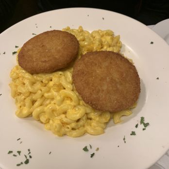 Alps Diner - A round white plate holds a serving of macaroni and cheese, with two breaded and fried items placed on top. The dish is garnished with small green herbs around the edge of the plate. The background is dark, and part of a small bowl of sauce is visible at the bottom right corner. - Diners Near Me - Wayne, New Jersey