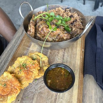 Travesias - Caribbean Oxtail - Oxtail stew with rice and cilantro garnish is served in a metal bowl, alongside fried plantains and a small container of sauce on a wooden board. - Latin American Near Me - Bronx, New York
