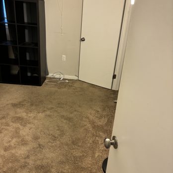 Reasonable Movers - A room with beige carpet, a white door slightly ajar, a black shelving unit, and a wall outlet with tangled cords are visible. - Movers Near Me