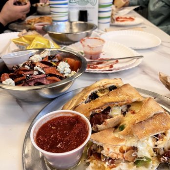 Blocky's Eatery - Hot and spicy calzone added sundried tomatoes!! Delicious don't skip on the garlic knots omg still thinking of them ! - Slices of pizza with cheese, meats, and vegetables are placed on a metal tray, accompanied by a cup of marinara sauce. The table has paper plates, cups, and a bowl of salad, with several people eating in the background. The setting appears to be a casual restaurant or pizzeria, with a sign for Blocky's Eatery visible in the background. - Italian Near Me - Loveland, Colorado
