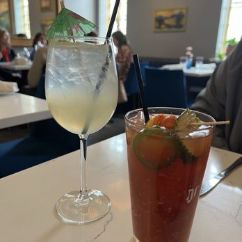 Mint - Limoncello Spritzer and Bloody Mary - Two cocktails, one with a small umbrella and a straw, are placed on a white table in a restaurant setting with patrons and artwork visible in the background. - Cocktail Bars Near Me - Glens Falls, New York